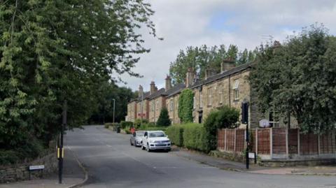 A road with houses on one side and a wall on the other side. Two cars parked in front of the houses. Trees on either side of the road.