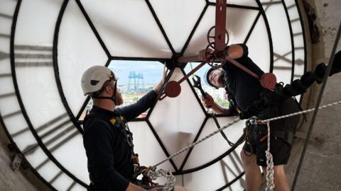 Joe Mines and Jason Budd are attached together with harnesses while removing the hands from a tower clock face. Both of the men are wearing safety helmets and Budd is leaning out of a hole in the glass face and holding a large wrench. The Transporter Bridge can be seen in the distance through a hole in the clock face.