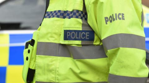 The torso of a police officer wearing a high-vis yellow jacket with the word Police written on it