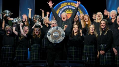 Men and women in the choir have their arms raised in celebration as they pose with trophies picked up during the competitions. The choir members are wearing dark tops and dark green tartan kilts.