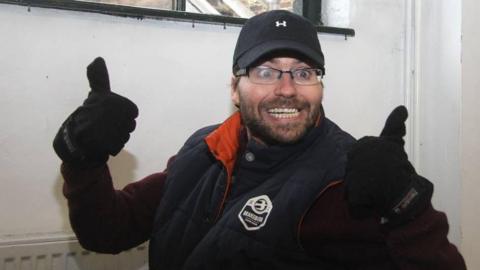 A man with a dark beard and moustache is giving a thumbs up signal and smiling. He is wearing a glasses, black cap, black gloves, dark jumper and dark sleeveless jacket. He is in front of a radiator.