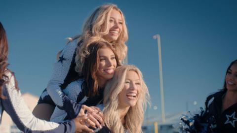 Pictured from top to bottom: Kleine, Lea, Megan. Kleine and Megan have long blonde hair and Lea has long brunette hair. They are shown jumping on each other while two squad members stand at the side