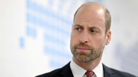Prince William, wearing a black suit, white shirt and red tie
