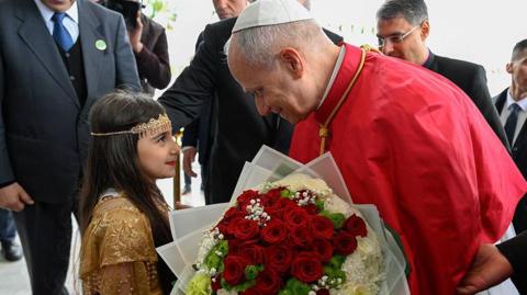 A young girl on the left wearing a gold sequinned dress and a beaded headdress. She is giving the pope - who is on the right and crouched down to her - a bouquet of red roses. He is smiling and wearing red. Partial shots of four men can be seen behind them.