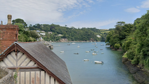 A peaceful river scene shows small boats anchored on calm water, with a traditional house and chimney in the foreground and tree-covered hills and scattered homes in the background.
