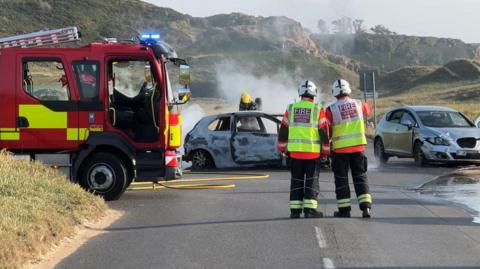Two fire engine parked on the left next to a car with smoke coming from it. There is a firefighter near the car, and two firefighters stood looking at the car. There's also another car to the right with damage to the front.