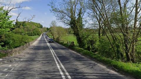 A street view image of a road winding its way through the countryside. Three cars can be seen travelling along the right lane, with trees and shrubs running along either side of the road.