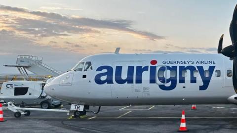 The image shows an Aurigny passenger plane parked on an airport tarmac, with the large blue word “aurigny” and the phrase “Guernsey’s Airline” visible on the side of the aircraft. Ground equipment and a small set of stairs are positioned next to the plane, with traffic cones placed around the area under a cloudy sky.