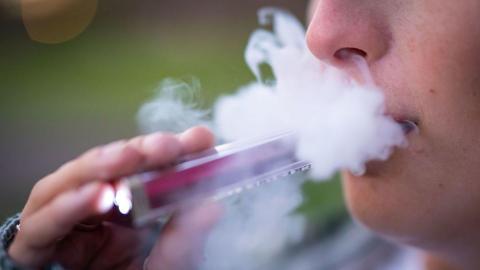 A stock image showing a young boy using a disposable pink vape, with a cloud of vapour going up his nose and out of his mouth.