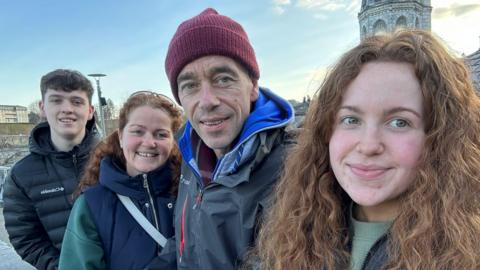 (L-R) Tiarnán, Debbie, John and Erin Gallagher are standing side by side. 
Tiarnán has black hair and is wearing a black hooded jacket.
Debbie us wearing a navy and green jacket.
John is wearing a red hat and blue hooded jacket, while Erin, who has long curly brown hair, is wearing a green jumper and jacket. 