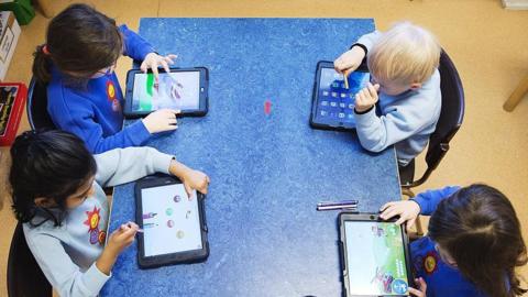 A group of children in a classroom. They all have their own tablet device and are wearing a blue school uniform