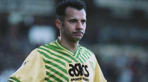 Dale Gordon is standing with his hands on his hips and is wearing a yellow and green Norwich City football jersey, branded with the words: Asics sports shoes. He is looking pensive. The background is blurred.