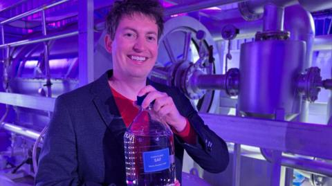 Bertie Stephens, CEO, Clean Planet Group, holding a bottle of Sustainable Aviation Fuel. Bertie, who is wearing a red top and suit jacket, is at the pilot facility in Sandwich, Kent. He has his left hand on the bottle top. He is smiling to camera.