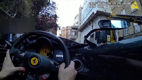 View from inside a Ferrari showing driver’s hands on the steering wheel in an urban area with construction scaffolding outside.