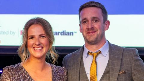 Head and shoulders image of Lizzie Walker and her husband at the awards ceremony. She has blond hair tied up and a sparkly dress and he is wearing a checked grey suit and gold tie