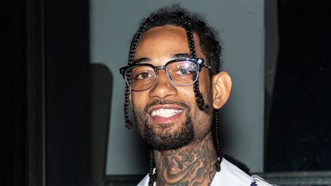 Rapper PnB Rock, in New York in 2020, wearing glasses with a black rim. Smiling. He is wearing a white top, and has tattoos on his neck. The background is of a window.