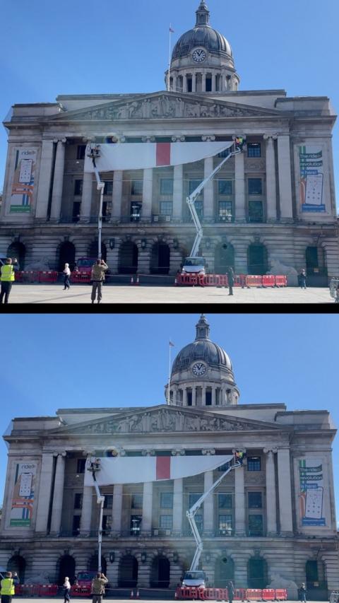 A large white flag with a red cross being unfurled on the front of a grand building with a domed clocktower.