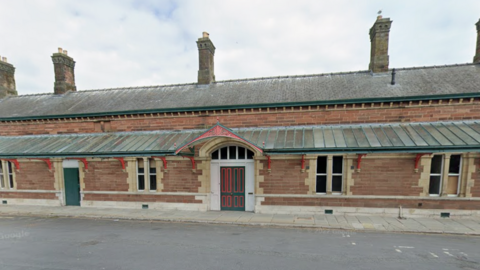 The brick-built front of Ulverston station with an empty road in front. The single-storey station has multiple chimneys, windows and doors. It has a lower roof and a second angled roof behind. It is a long building. It has a door painted green and red in the middle. 
