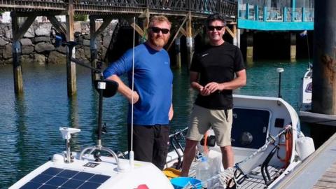 Tim Foster and Mike Reed stand on their rowing boat, Cheerfulness in dock. It is white and has solar panels on the front along with a Manx flag sticker. Both men wear sunglasses and are middle aged, with ginger and dark hair.