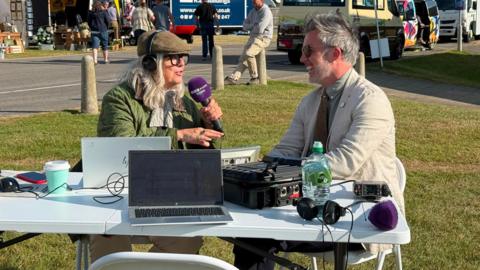 A picture of a radio presenter interviewing a guest at The Great Yorkshire Show. The presenter is wearing a flat cap, a green jacket and is holding a microphone. The guest is laughing and wearing a pale jacket. They are sitting at a table in front of an ice cream van.