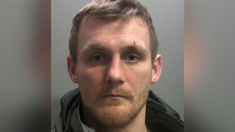 A mugshot of Marc Proudfoot, a man in his 30s with short hair and stubble. He appears to have a marking above one of his eyebrows - it is not clear whether it is a wound.