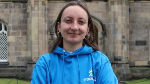 Christina Schmid, a woman with long brown hair, a blue hoodie and nose piercings, looks into the camera