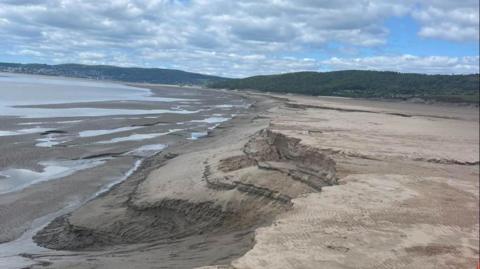 It is a cloudy day over Morecambe Bay with some blue patches of sky. There are patches of water along the bay with steep drops evident in parts of the sand. 