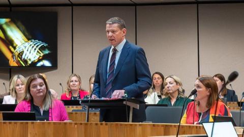 A man, Rhun ap Iorwerth, is in a navy suit and is standing up in the Senedd chamber surrounded by other seated members.