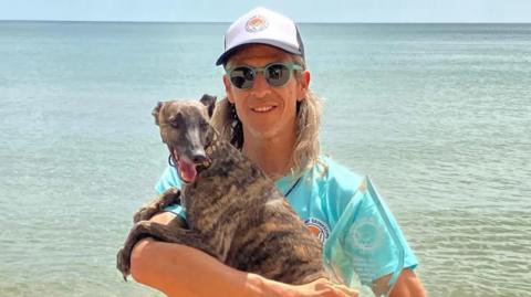 Man in a green t-shirt standing in front of the sea, holding a whippet in his arms