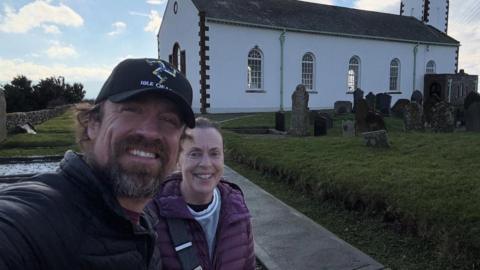Reed Timmer on the left, has dark hair and a beard and is wearing a black coat and black cap with the Three Legs of Man emblem on it. Next to him to the right is his mum smiling, wearing a purple jacket and her dark hair tied back. They're standing in a churchyard with grave stones and a white church building behind them to the right on a sunny day.