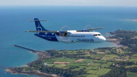 A Blue Islands plane flies over Jersey on a bright day. The sky is blue and the land below is patchworked with fields and has beaches surrounding it. There is a large quay visible.