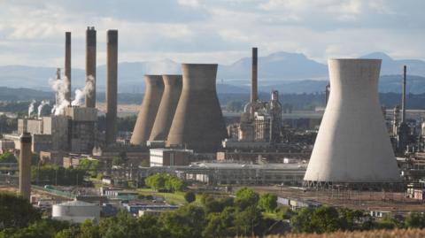 A view of the Grangemouth petrochemical plant in Grangemouth. A large white tower is in the foreground, with three darker ones further away. Smoke billows from the chimneys f various buildings in the heavily industrial site