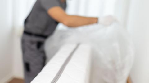 A service worker is removing the plastic covering from a mattress in a home. The room has light-coloured walls and wooden flooring. The activity takes place during the daytime. This is a stock image.