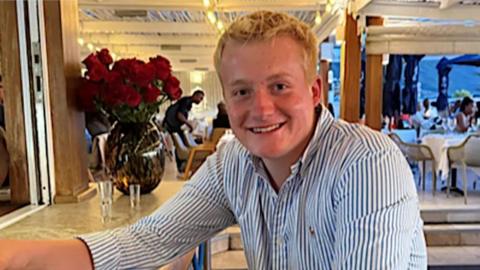 Harley Pearce sits at a bar in a blue and white striped shirt. He appears to be in a relatively fancy restaurant or hotel, with tables visible behind him. He has short, blond hair and is smiling at the camera.
