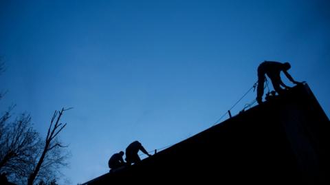 Silhouetted people work on top of a roof.