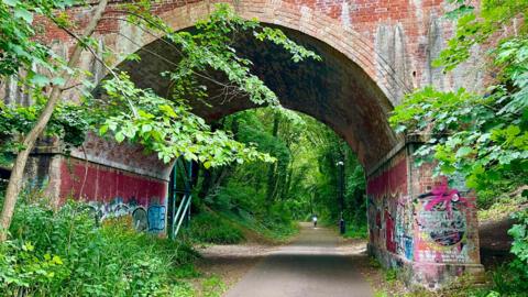 A brick bridge covered in graffiti with a path going through the centre. There is a black lamp post. There are green trees either side of the path.
