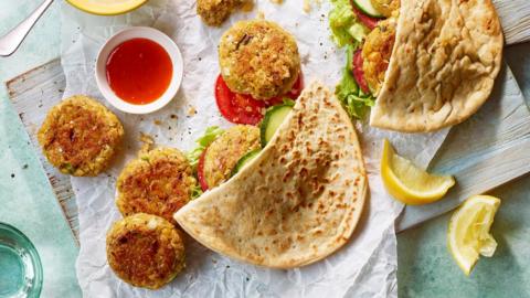 A halved pitta bread on a board with rounds of homemade falafel and salad spilling out, some squeezed lemon wedges and a bowl of chilli sauce