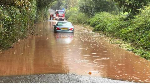 A silver vehicle in the middle of a flooded road heading into Crediton from Sandford. The wheels of the car are under the brown coloured flood water. In the background is a red fire engine with its blue lights on. To the right of the fire engine are crews. 