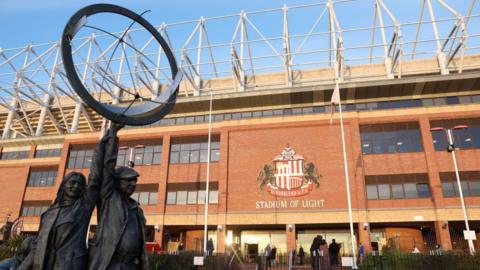 A general view of the Stadium of Light in Sunderland. It is made of brick with multiple windows. It has a Stadium of Light sign and a statue of a man and a woman stand in the foreground holding a circle shape.