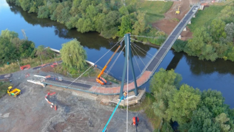 An aerial view of a bridge, midway through construction, over a river with trees on both banks and construction vehicles in the foreground.