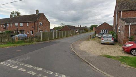 A street view shows a juntion at a road. There are redbrick houses and cars parked on driveways