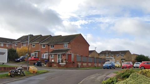 Mill Green Road on an overcast day. The houses are made with red bricks and there are cars parked along the road.