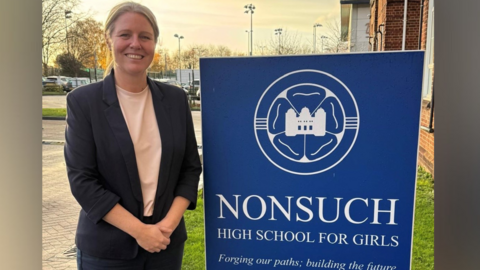 Alexis Williamson, the head teacher of Nonsuch High School for Girls. Alexis is standing beside a blue sign for the school. She has her right hand over her left hand, and is smiling to camera. 