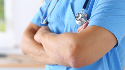 A hospital worker, wearing blue scrubs, with their arms folded. They have a stethoscope around their neck. 