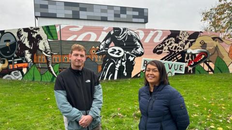 A man and a woman stand side by side in front of a colourful mural in a park. 