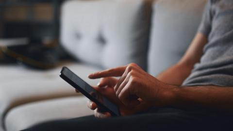 A stock image of a man using a smartphone while sat on a grey sofa