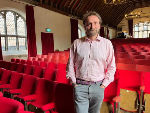 Tim FitzHigham stands inside St George's Guildhall. The roof is made up of timber beams. Below pointing towards the stage are rows of red seats for the audience. 