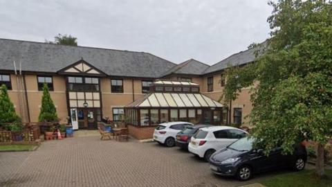 The former Hallgarth Care Home is a two-storey beige brick building with a small outside seating area and a four parked cars.