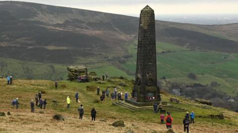 Scores of people gather while socially distancing during a memorial event in 2020 around the Saddleworth memorial. It is a tall slender obelisk pillar on a hill, surrounded by other hills in the Pennine countryside.