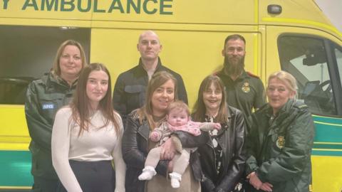 Four ambulance paramedics (two male, two female) and two woman, one in a white top and one in a black top, stand in front of an ambulance. The woman in a black top is holding a baby.